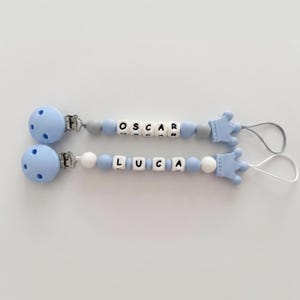 May include: Two light blue dummy clips featuring silicone beads in shades of blue, white, and grey. Each clip includes white cube beads spelling the names OSCAR and LUCA, a metal attachment clip, and a light blue silicone crown-shaped end piece.