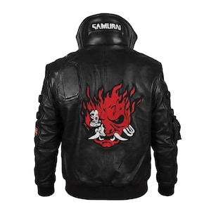 Op de afbeelding: Een zwart leren bomberjack met een grote rood-witte afbeelding van een gestileerd oni-masker op de achterkant. De kraag toont het woord SAMURAI in witte letters. Het jack heeft geribbelde boorden, een hoge kraag en diverse textuurpanelen.