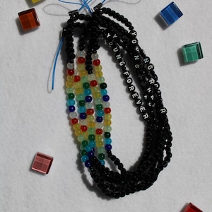 May include: A collection of beaded phone charms in black and rainbow colors. One charm features the text "YOUNG FOREVER" in white beads. Scattered around are colorful, square-shaped glass beads.