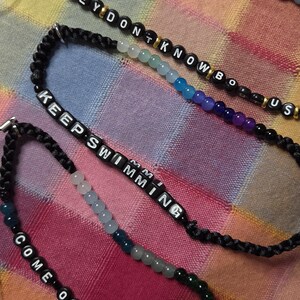 May include: Three adjustable black braided cord bracelets featuring colorful beads and lettered cubes. The top bracelet reads THEY DONT KNOW US, the middle reads KEEP SWIMMING, and the bottom reads COME OVER, all accented with gold-toned spacers.