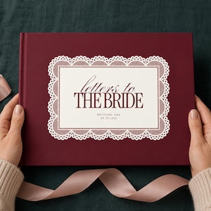 May include: A deep burgundy hardcover book with a cream-colored rectangular center panel featuring a white lace-style border. The text reads "letters to THE BRIDE" with "BRITTANY ANA" and "09.19.2026" printed in dark burgundy below the title.