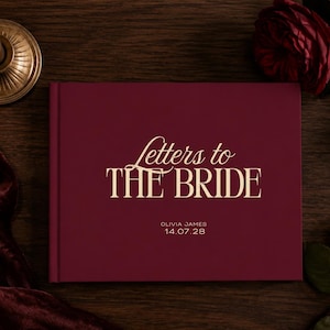 Könnte beinhalten: Ein tiefburgunderrotes Hardcover-Buch mit goldener Folienprägung, die Letters to THE BRIDE sowie den Namen OLIVIA JAMES und das Datum 14.07.28 zeigt. Das Buch liegt horizontal auf einer dunklen Holzoberfläche mit einer Kerze und einer Rose.