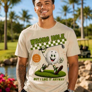 May include: Beige t-shirt with the text "WRONG HOLE" above a cartoon golf ball character holding a golf club. The golf ball is on a patch of green grass, with the text "BUT I LIKE IT ANYWAY" below.