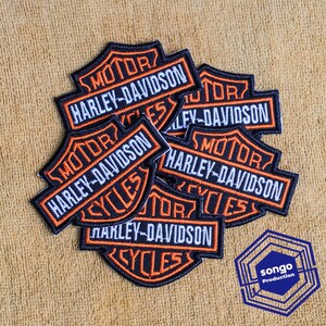 Op de afbeelding: Een verzameling geborduurde Harley-Davidson Motor Cycles patches met een schildvormig ontwerp in zwart, wit en oranje garen. Elke patch toont de tekst MOTOR, HARLEY-DAVIDSON en CYCLES in een vetgedrukte, duidelijke letterstijl.