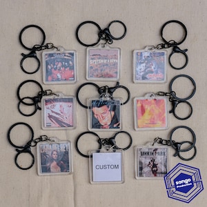 Könnte beinhalten: Eine Sammlung von neun quadratischen Acryl-Schlüsselanhängern mit verschiedenen Musikalbum-Cover-Drucken und einem weißen Quadrat mit dem schwarzen Text CUSTOM. Jeder Schlüsselanhänger ist an einem schwarzen Metallring und einem kleinen Verbindungsring befestigt.