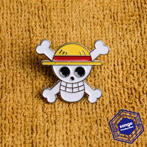 Puede incluir: Un pin de esmalte metálico que muestra una calavera blanca con grandes ojos negros y una mandíbula con patrón de cuadrícula, usando un sombrero de paja amarillo con una banda roja. Dos huesos blancos cruzados se sitúan tras la calavera.
