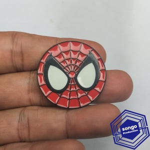 May include: A circular enamel pin featuring a red and black spiderweb pattern with two large white eye shapes in the centre. The design is inspired by a comic book character mask, rendered in bold colours with a glossy finish and metallic outlines.