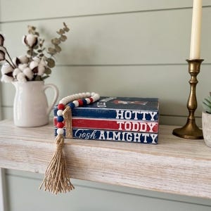 May include: A stack of three rectangular wooden blocks painted in navy blue, red, and navy blue, featuring the white text Hotty Toddy Gosh Almighty. A strand of red, white, and blue wooden beads with a jute tassel is draped over the stack.