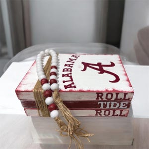 May include: A white and red wooden box with the word "ALABAMA" and the letter "A" in crimson. The box is wrapped with twine and adorned with a beaded garland of white and crimson beads. The words "ROLL TIDE ROLL" are printed on the side.