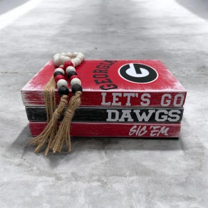 Puede incluir: Una pila de tres libros decorativos de madera en rojo, negro y blanco. El libro superior es rojo con la palabra "GEORGIA" y un logotipo. El libro del medio es negro con "LET'S GO DAWGS" en blanco. El libro inferior es rojo con "SIC 'EM" en blanco. Una guirnalda de cuentas rojas, negras y blancas descansa encima.