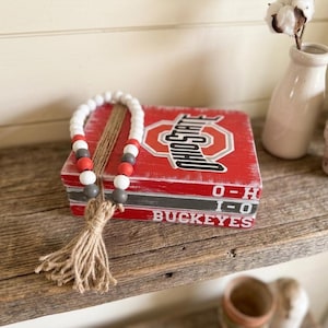 May include: A stack of three rectangular wooden blocks painted red, white, and gray with the text OHIO STATE, O-H, I-O, and BUCKEYES. A strand of white, red, and gray wooden beads with a jute tassel is draped over the top of the block stack.