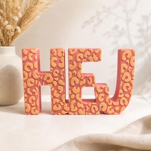 Könnte beinhalten: Eine korallenfarbene dekorative Skulptur mit der Aufschrift "HEJ" in Großbuchstaben. Die Buchstaben sind mit einem beigefarbenen Leopardenmuster verziert. Die Skulptur wird auf einer weißen Oberfläche ausgestellt, mit einer Vase mit getrockneten Pflanzen im Hintergrund.