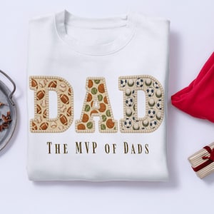 Könnte beinhalten: Weißes Sweatshirt mit dem Wort "DAD" in großen, strukturierten Buchstaben, die mit Sportbällen gefüllt sind. Der Text "THE MVP OF DADS" ist darunter gedruckt. Das Sweatshirt ist auf einer weißen Oberfläche gefaltet.