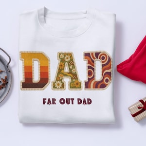 May include: White sweatshirt with the word "DAD" in large, colorful, retro-style lettering. The "D" is striped, the "A" has a floral pattern, and the second "D" has a swirl design. Below, the text reads "FAR OUT DAD".