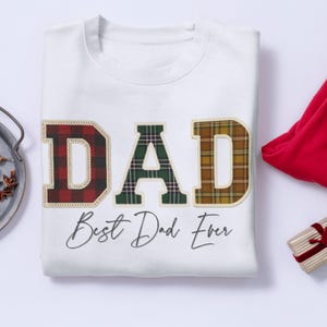 Puede incluir: Sudadera blanca con la palabra "DAD" en letras grandes a cuadros. Las letras son rojas, verdes y doradas. Debajo de la palabra "DAD" está la frase "Best Dad Ever" en cursiva.