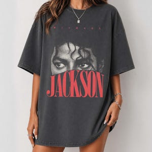 May include: Dark gray oversized t-shirt featuring a graphic of Michael Jackson's eyes and the name "JACKSON" in red block letters. The word "MICHAEL" is printed above the eyes in red. The shirt has short sleeves and a relaxed fit.