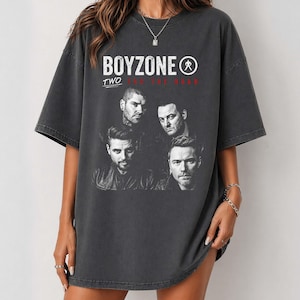 May include: A charcoal gray short-sleeve t-shirt featuring a black and white graphic print of the band Boyzone. The text reads "BOYZONE" in large white letters above "TWO FOR THE ROAD" in smaller red text, with a circular logo icon included.