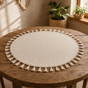 Puede incluir: Un mantel individual circular de tela color blanco roto con un borde decorativo de ganchillo adornado con numerosas borlas pequeñas que cuelgan por todo el perímetro. El mantel tiene un centro liso y liso sobre una superficie de madera.