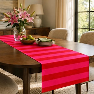 May include: A rectangular table runner featuring alternating horizontal stripes in vibrant shades of hot pink and bright red. The fabric runner is laid flat across the centre of a dark wooden dining table, providing a bold colour contrast.