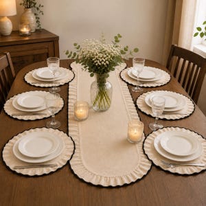 May include: A beige table runner with ruffled edges and a black trim, centred on a wooden table. The setting includes four round placemats with matching ruffled edges, white plates, clear glass goblets, two lit candles, and a glass vase of flowers.