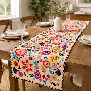 May include: A cream-coloured table runner featuring a vibrant, multi-coloured floral pattern with red, yellow, pink, and purple blossoms. The runner has a scalloped edge with a black trim and is laid across a wooden dining table with place settings.