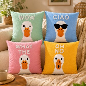Puede incluir: Cuatro cojines cuadrados con caras de pato y texto. Los cojines son en tonos de verde, azul, rosa y amarillo. Cada cojín tiene una cara de pato blanca con un pico naranja. El texto en los cojines dice "WOW", "CIAO", "WHAT THE" y "OH NO".