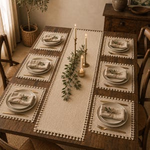 May include: A rectangular beige table runner and six matching placemats, all featuring decorative fringe trim. Each place setting includes a ceramic plate, a folded linen napkin tied with twine and a small green leaf, and gold-toned cutlery.