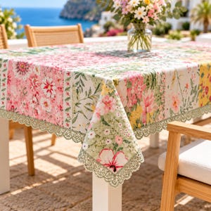 Puede incluir: Un mantel rectangular con un diseño de patchwork de varios patrones florales en tonos rosa, verde, amarillo y blanco. La tela está rematada con un borde decorativo de encaje festoneado a lo largo de los lados, cubriendo una mesa.