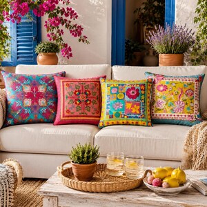 May include: Four decorative throw pillows with colorful floral patterns rest on a white sofa. The pillows feature vibrant hues of pink, turquoise, yellow, and green. The scene is set outdoors with plants and flowers.