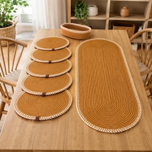 May include: A set of mustard yellow and white rope-coiled table linens, including one large oval runner, five oval placemats, and one small oval basket. Each piece features a continuous spiral design with a thick, braided white border trim.