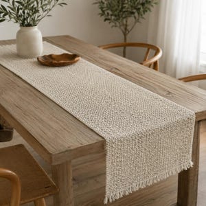 Puede incluir: Un camino de mesa rectangular de color beige con un patrón de tejido texturizado y flecos cortos en los extremos. El camino está colocado plano sobre el centro de una mesa de comedor de madera, proporcionando una base neutra.