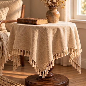 May include: A round beige linen tablecloth with a textured weave and decorative fringe tassels along the edges. The fabric drapes over a circular wooden pedestal table, creating soft folds. The material has a natural, neutral tan color.