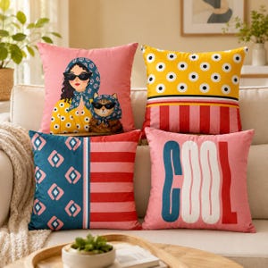 May include: A collection of decorative throw pillows in various designs. One pillow features a woman and a cat wearing sunglasses and headscarves. Other pillows have geometric patterns and the word "COOL". The color palette includes pink, yellow, blue, and red.
