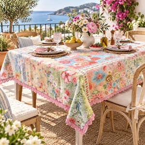 Puede incluir: Un mantel rectangular con un vibrante estampado floral en tonos rosa, verde y amarillo, rematado con un delicado borde de encaje de ganchillo rosa. La tela presenta un diseño botánico repetitivo con diversas flores en toda su superficie.