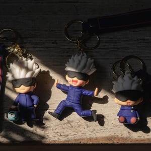 Op de afbeelding: Drie sleutelhangers in anime-stijl met figuren met wit of zwart stekelig haar, zwarte blinddoeken en donkerblauwe uniformen. Elke sleutelhanger bevat een metalen ring en een lange, donkerblauwe stoffen band met een rode rand.