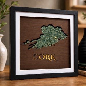 Może przedstawiać: Oprawiona dekoracja ścienna z ciemnym drewnianym tłem i wyciętą mapą hrabstwa Cork w Irlandii. Mapa ma zieloną powierzchnię z misternymi złotymi liniami. Słowo CORK jest wycięte poniżej w formie złotych liter.