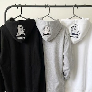 Puede incluir: Tres sudaderas con capucha en negro, gris y blanco colgadas en un perchero. Cada capucha presenta un gráfico de un fantasma haciendo un gesto obsceno. La sudadera negra dice FUCK U, la gris FUCK U BOTH y la blanca FUCK U 2.