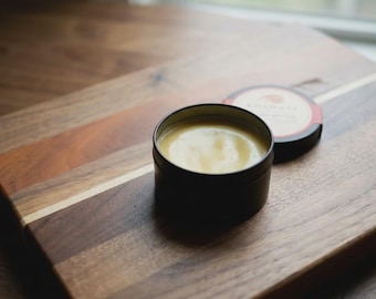 May include: A small, round, black metal tin filled with a smooth, pale yellow balm. The lid is placed beside the open container, featuring a circular label with a red border and a graphic of a sun, with text that is partially visible.