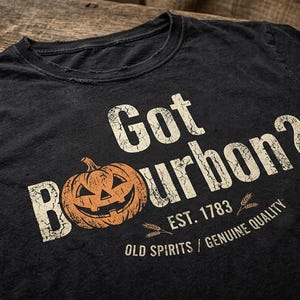 May include: A black short-sleeved t-shirt featuring a distressed white graphic that reads Got Bourbon? with an orange jack-o'-lantern replacing the letter O. Below the text is the phrase EST. 1783, wheat stalks, and OLD SPIRITS / GENUINE QUALITY.