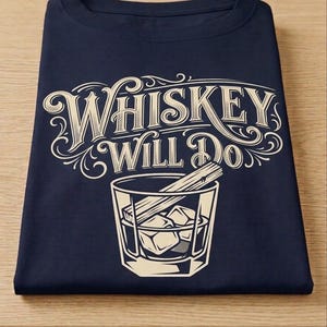 May include: A navy blue t-shirt featuring a cream-colored graphic of a glass filled with ice cubes and a wooden stirrer. Above the glass, the text reads "WHISKEY WILL DO" in a decorative, vintage-inspired font with ornate flourishes.