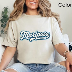 May include: A sand-colored short-sleeve t-shirt featuring the word Mariposa in a white script font with a dark blue and light blue outline, and the word Monarchs in smaller white text underneath, paired with light blue distressed denim jeans.