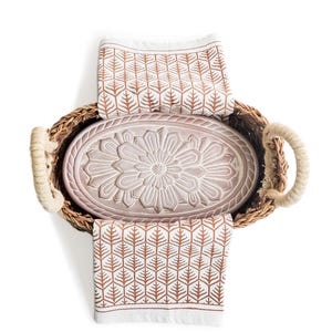 May include: An oval woven basket with thick rope handles containing a light pink ceramic tray with a central floral relief pattern. Two white fabric cloths with a repeating rust-colored geometric tree-like print are draped over the basket.
