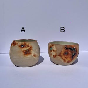 以下が含まれることがあります： AとBのラベルが付いた2つの陶器製カップ。カップAは背が高く丸みを帯びており、カップBは背が低く幅広です。どちらも明るいベージュのベースで、外側には焦げたオレンジ、茶色、濃い灰色の質感のある斑点模様が施されています。