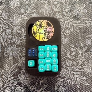May include: A black 3D-printed organizer tray containing a collection of gaming dice. It features a circular sticker with a yellow and orange dragon illustration, one translucent blue six-sided die, and nine turquoise dice marked with numbers.