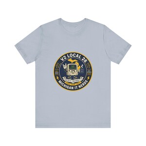 May include: A light blue t-shirt featuring a circular graphic on the chest. The design includes a cartoon computer character, the text V2 LOCAL 26, MICHIGAN IT NERDS, EST. 2024, WE FIX IT, YOU ROCK IT, and a map of Michigan in yellow and dark blue.