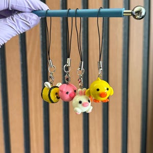 Puede incluir: Una colección de cinco colgantes de animales coloridos, incluyendo una abeja, un cerdo, un conejo y un pato, suspendidos de una barra turquesa. Cada colgante está unido a un cordón negro con un cierre plateado. La abeja es amarilla y negra, el cerdo es rosa.