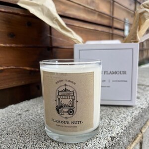 Op de afbeelding: Een heldere glazen kaars met crèmekleurige was en een bruin etiket. Op het etiket staat "MAISON FLAMOUR" en "FLAMOUR NUIT. Fabriqué en Espagne." Op de achtergrond staat een witte doos met "FLAMOUR".