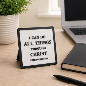May include: A square desk sign with a black frame and a white center panel featuring the black text I CAN DO ALL THINGS THROUGH CHRIST PHILIPPIANS 4:13. The sign is supported by a small black easel stand on a light wood surface.
