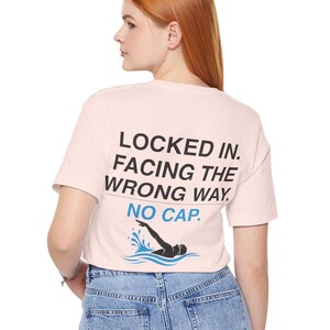 Puede incluir: Una camiseta rosa claro con el texto "LOCKED IN. FACING THE WRONG WAY. NO CAP." en negro. Debajo del texto hay un gráfico de un nadador en agua azul. La camiseta la lleva una persona con vaqueros azules.