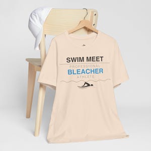 Könnte beinhalten: Ein hellbeige T-Shirt mit den Worten "SWIM MEET" über "PROFESSIONAL BLEACHER ATHLETE" in schwarzer und blauer Schrift. Eine Schwimmer-Grafik befindet sich unter dem Text. Eine weiße Baseballkappe liegt auf einem Holzstuhl.